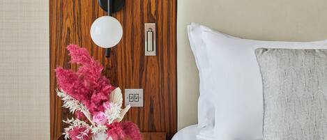 A neatly made hotel room with a wooden headboard, a bedside table with a lamp, a vase with pink flowers, and a bed with white and gray bedding.