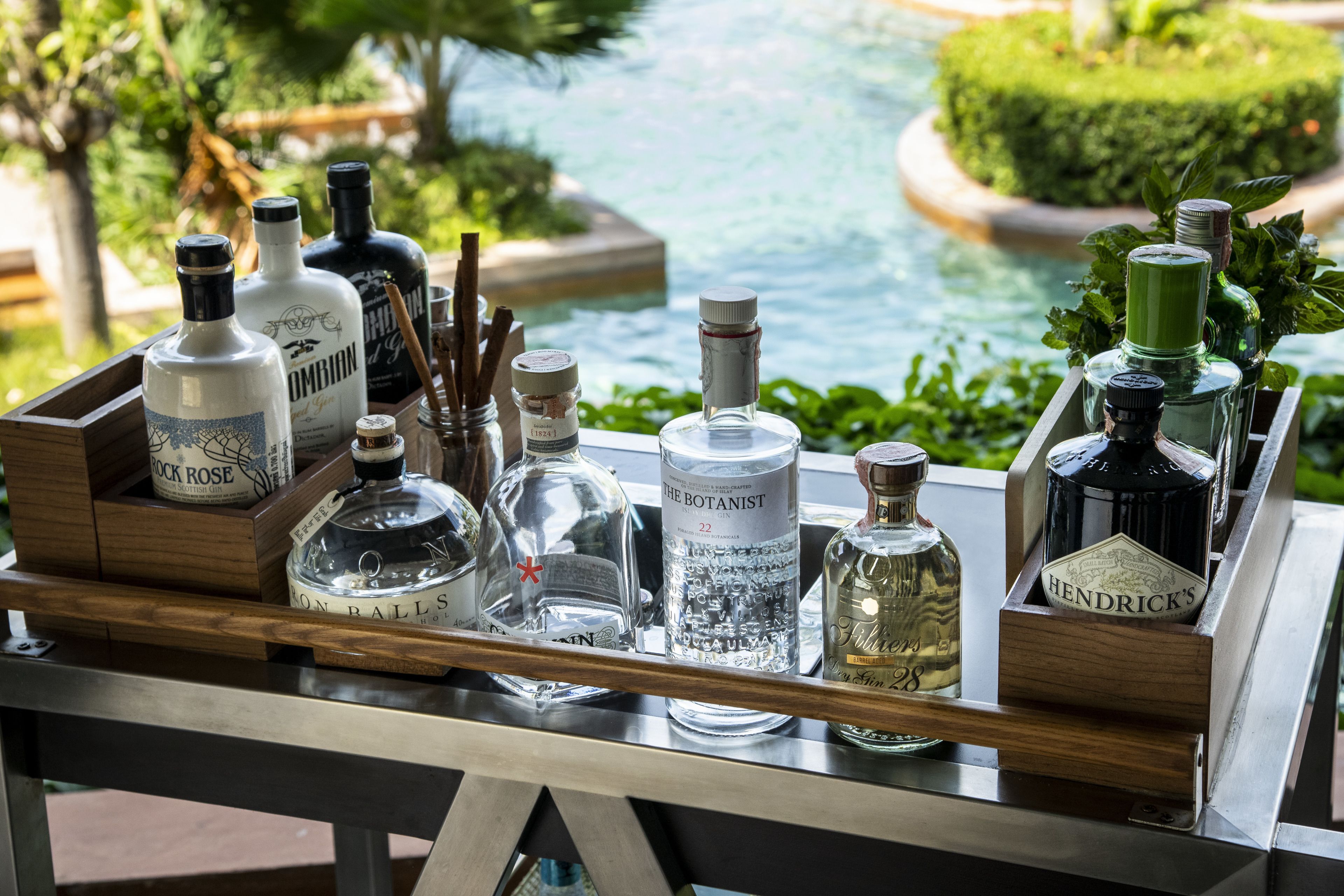 Un plateau en bois contenant différentes bouteilles d’alcool et un petit pot d’épices, posé sur une table d’où l’on peut admirer une piscine et la verdure environnante.