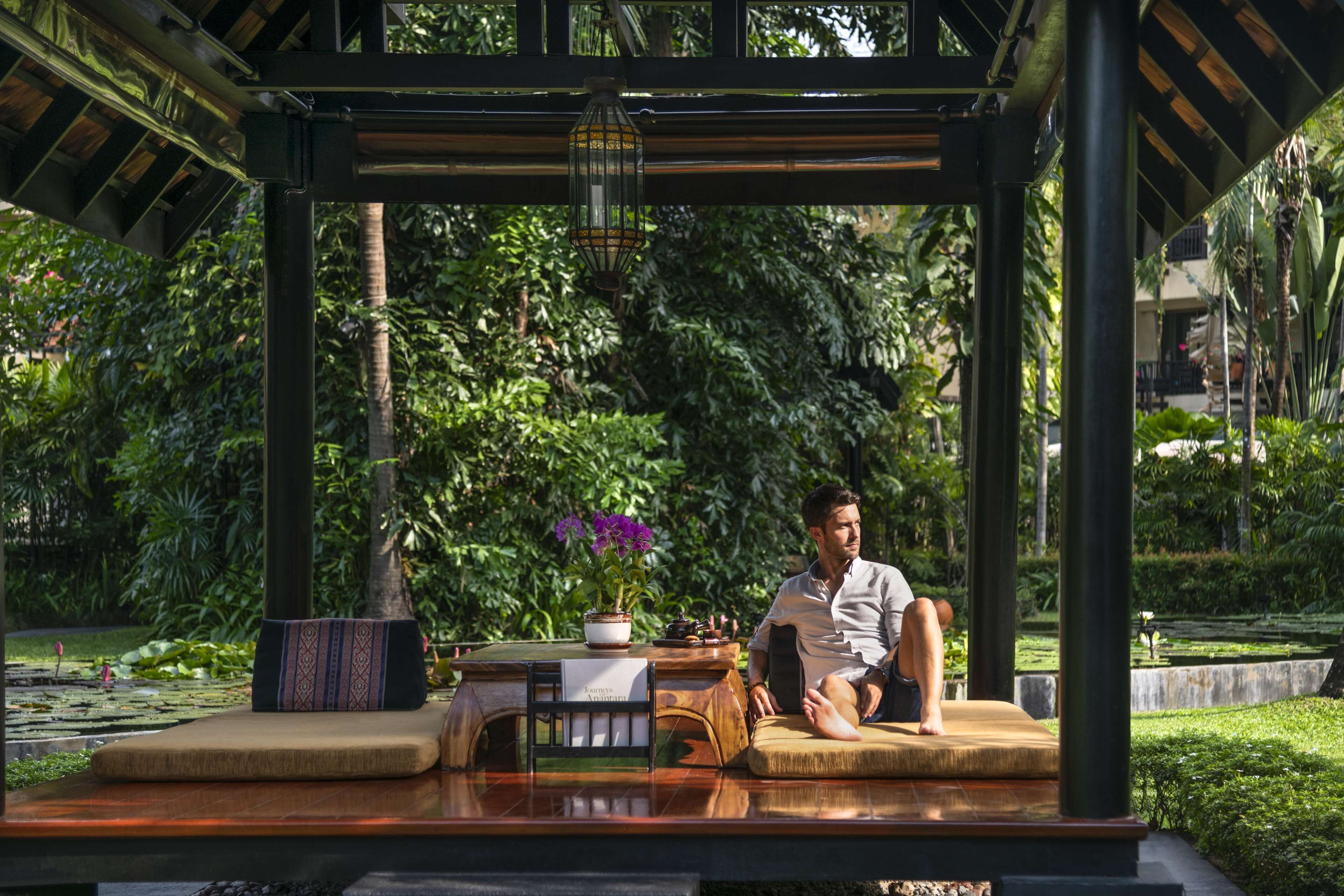 A man sitting on a wooden bench under a pergola with a potted plant and a lantern.