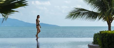 Une femme en bikini, debout au bord d’une piscine, avec la mer et les montagnes en toile de fond.
