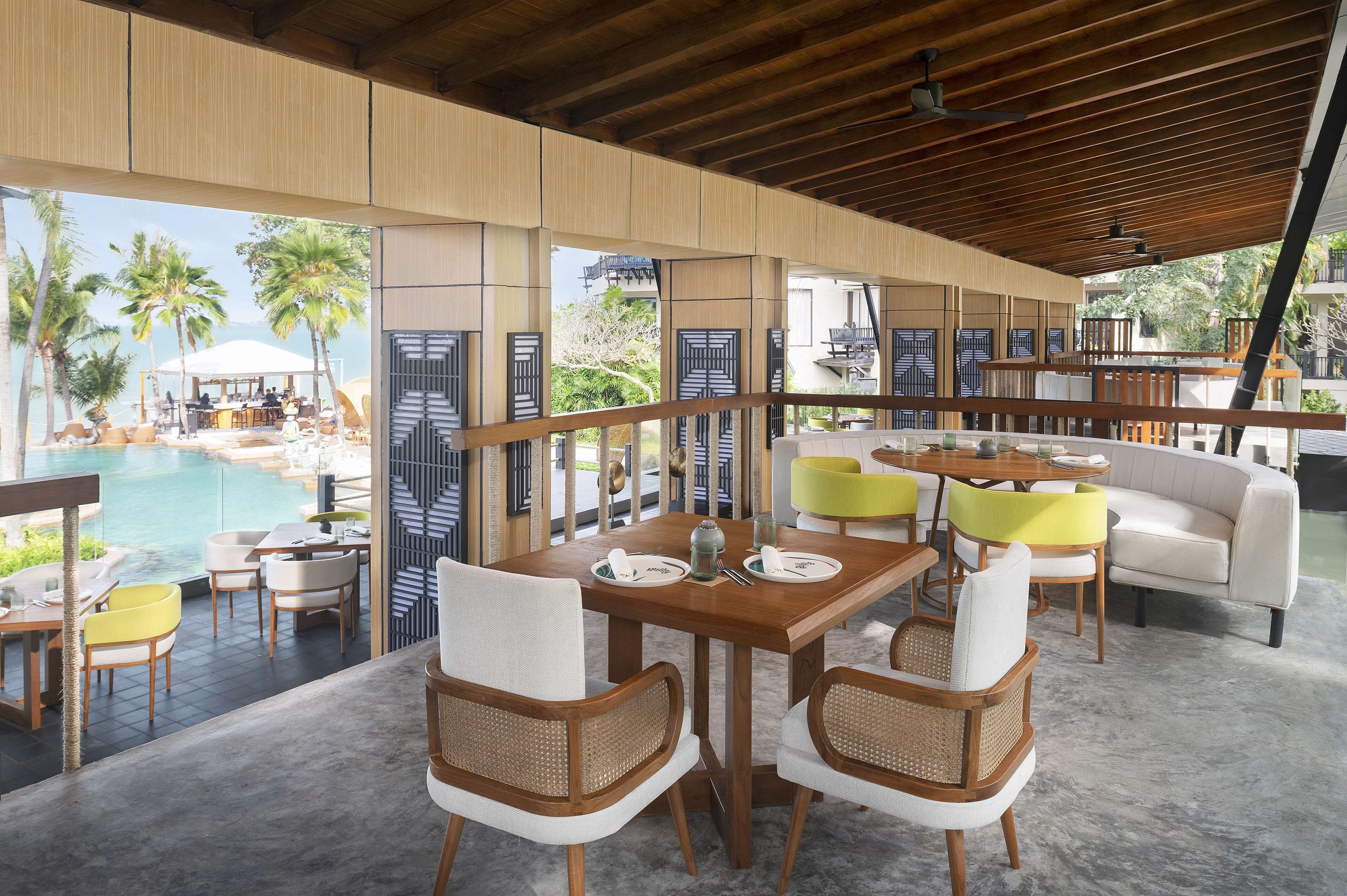 A modern outdoor dining area with a pool view, wooden furniture, and a tropical backdrop.