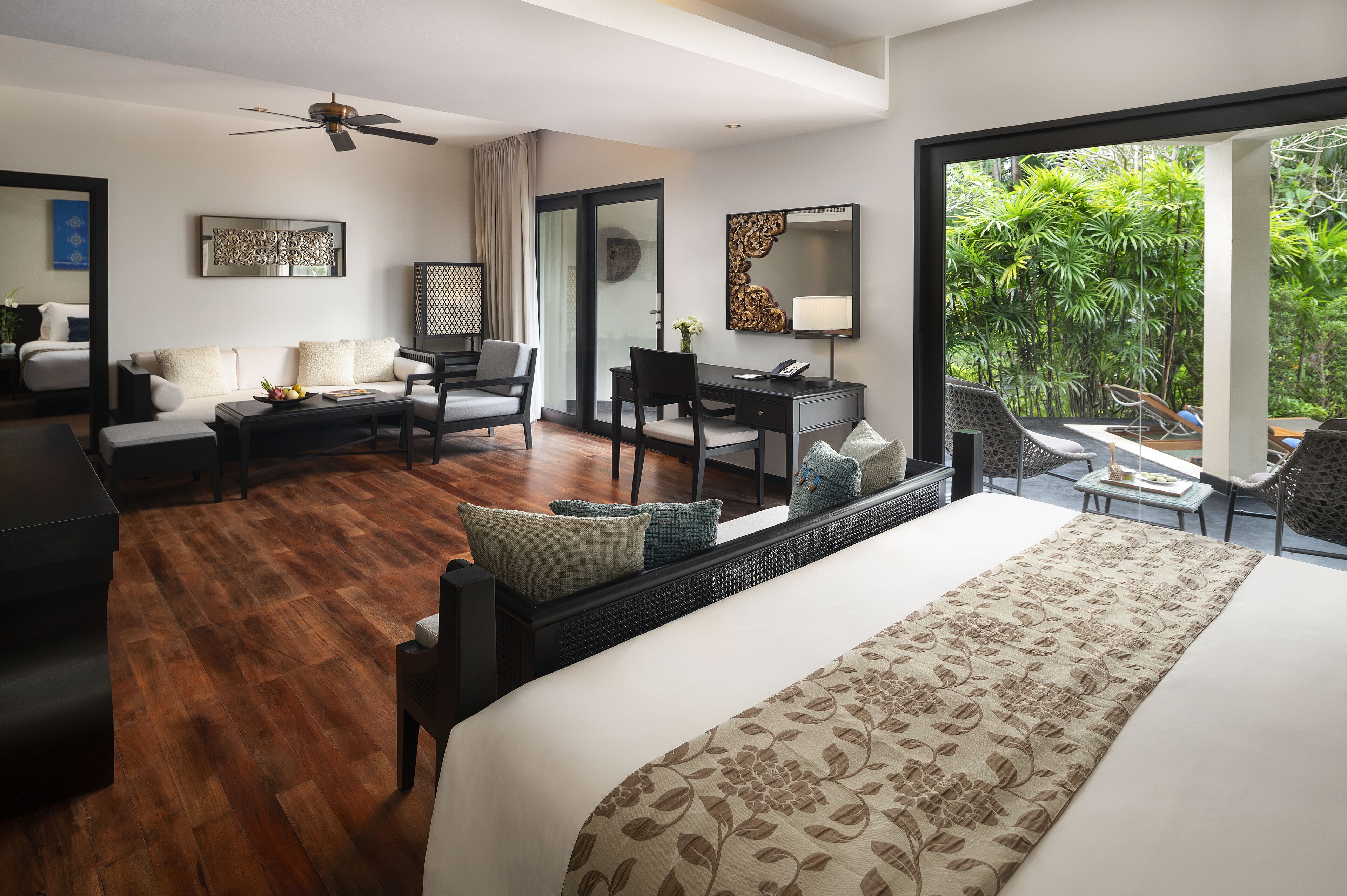 A modern hotel room with a large bed, a desk, a sofa, and a balcony with a view of greenery.