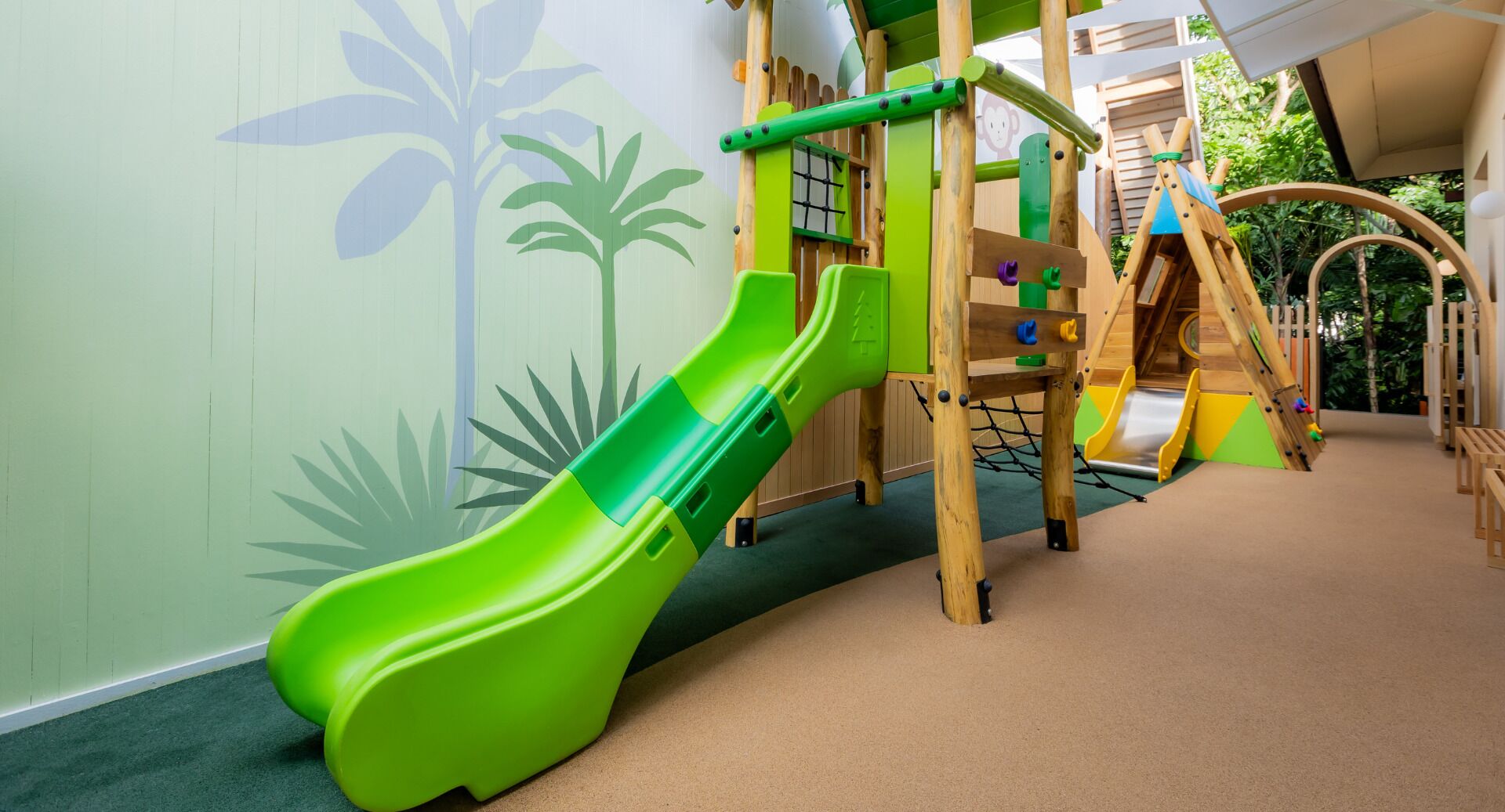 Un espace de jeux pour enfants avec un toboggan vert, un filet d’escalade et des structures de jeu en bois.