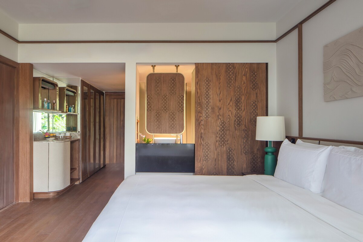 A hotel room with a large bed, a nightstand, a mirror, a wooden panel wall, and a bathroom visible through an open door.