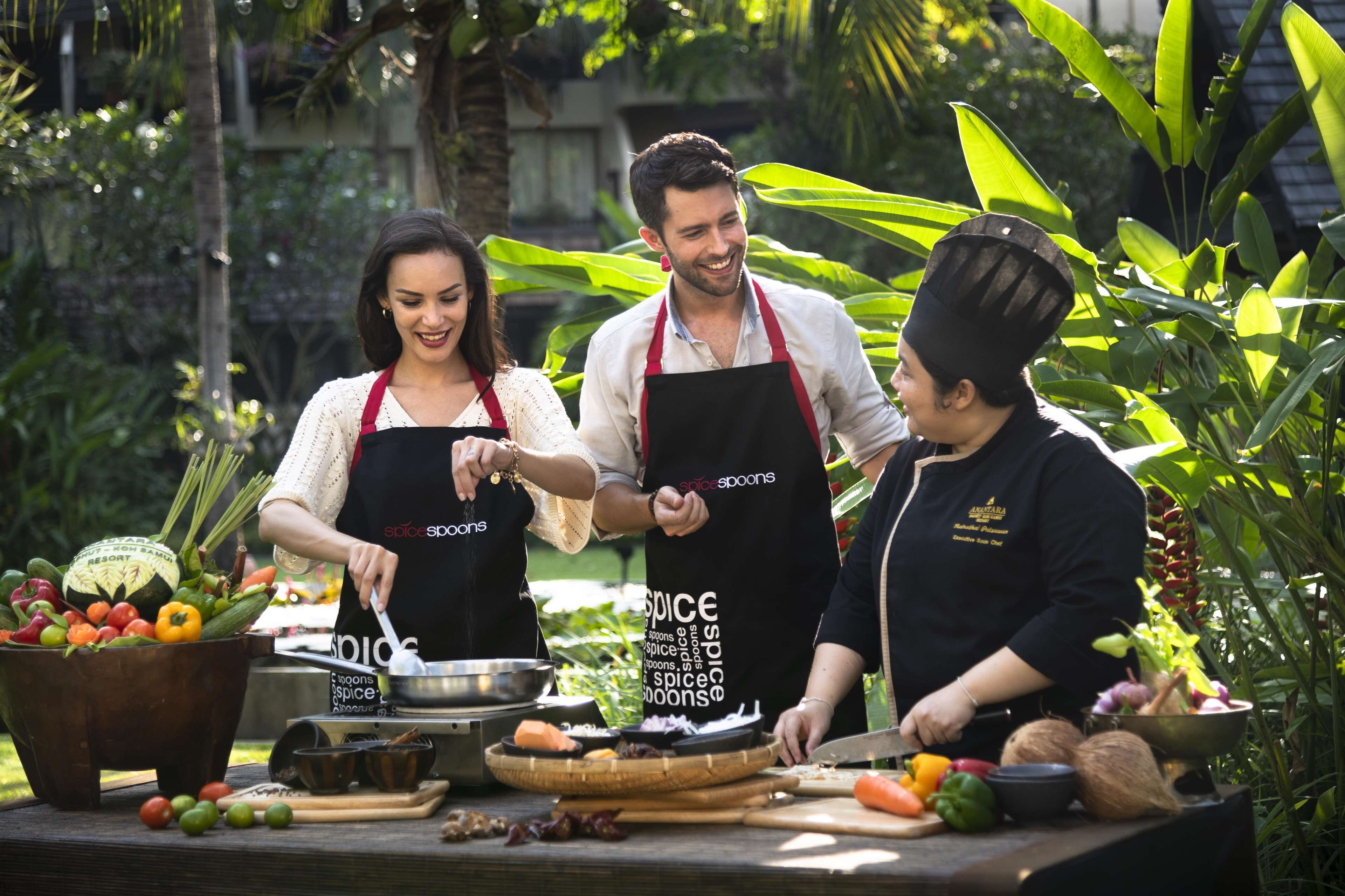 Trois personnes portant des tabliers arborant le logo « Spice Spoons », préparant des aliments à l’extérieur, avec une variété de légumes et d’ustensiles de cuisine visibles.