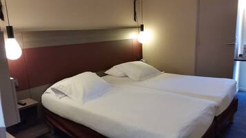 Une chambre d’hôtel avec deux lits, chacun étant équipé de draps et d’oreillers blancs. On y trouve deux lampes de chevet et une table de chevet avec une télécommande.