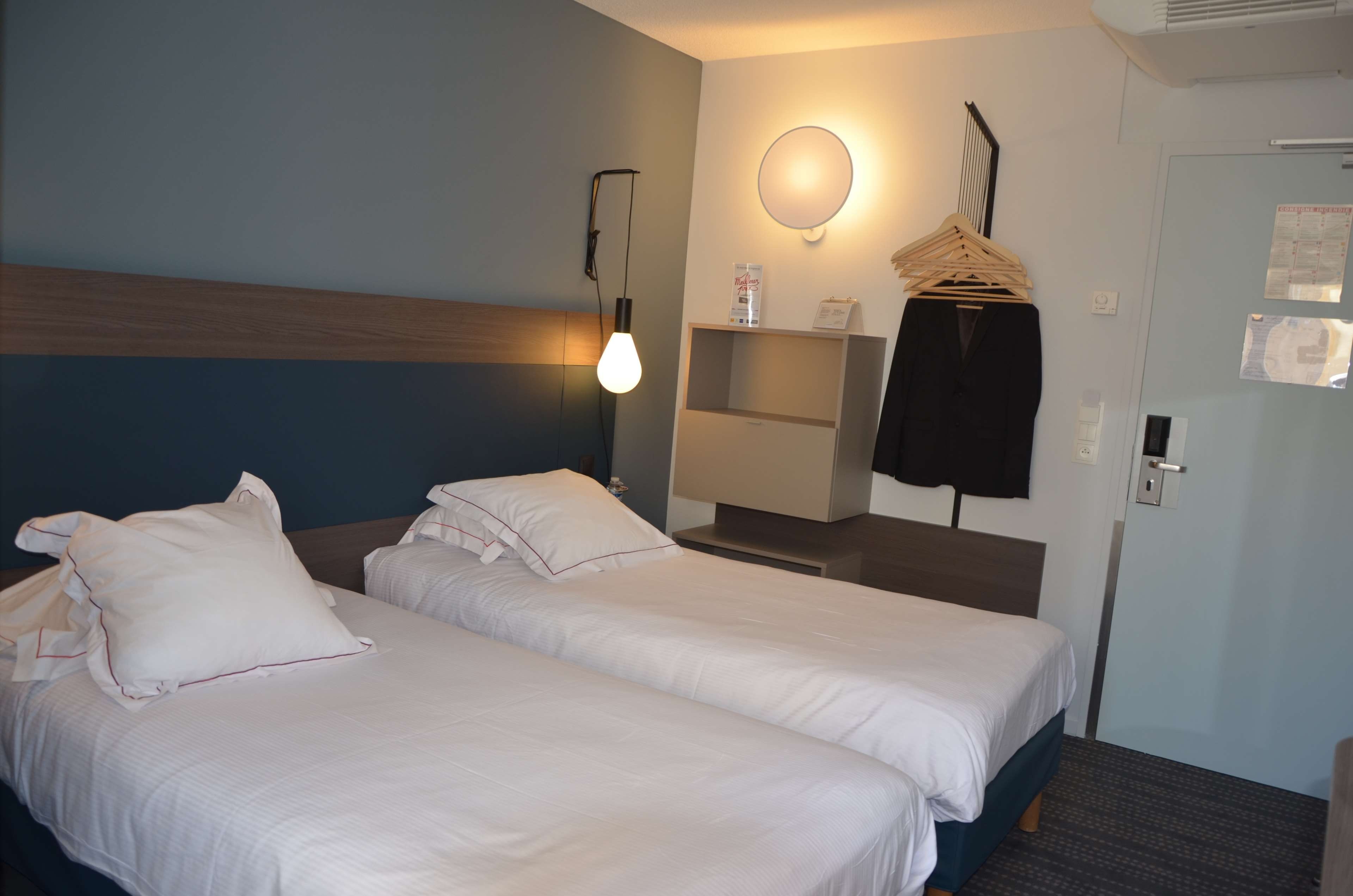 A hotel room with two beds, a bedside table, a mirror, a hanging coat rack, and a wall-mounted heater.