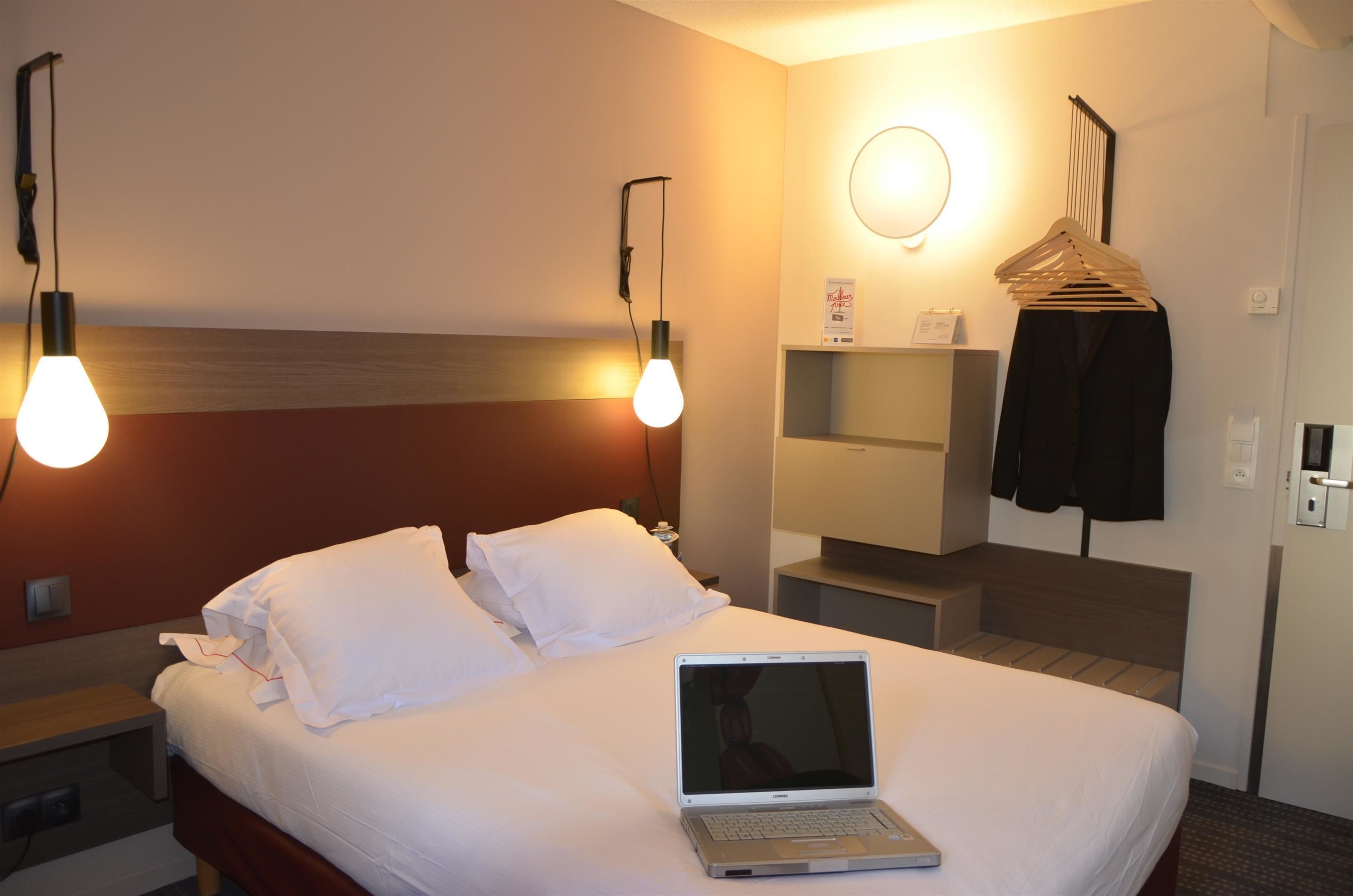 A hotel room with a bed, a laptop, a bedside table, a nightstand, a wall-mounted shelf, a mirror, and a coat rack with hangers.