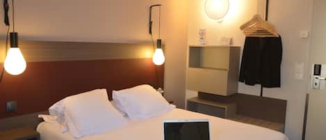 Une chambre d’hôtel comprenant un lit, un ordinateur portable, une table de chevet, une commode, une étagère murale, un miroir et un porte-manteau avec des cintres.