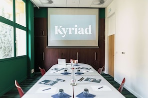 Kokoushuone, jossa on pitkä pöytä, tuoleja, projektorikangas, jossa näkyy teksti ”Kyriad”, ja ikkuna, josta avautuu näkymä puita kohti.