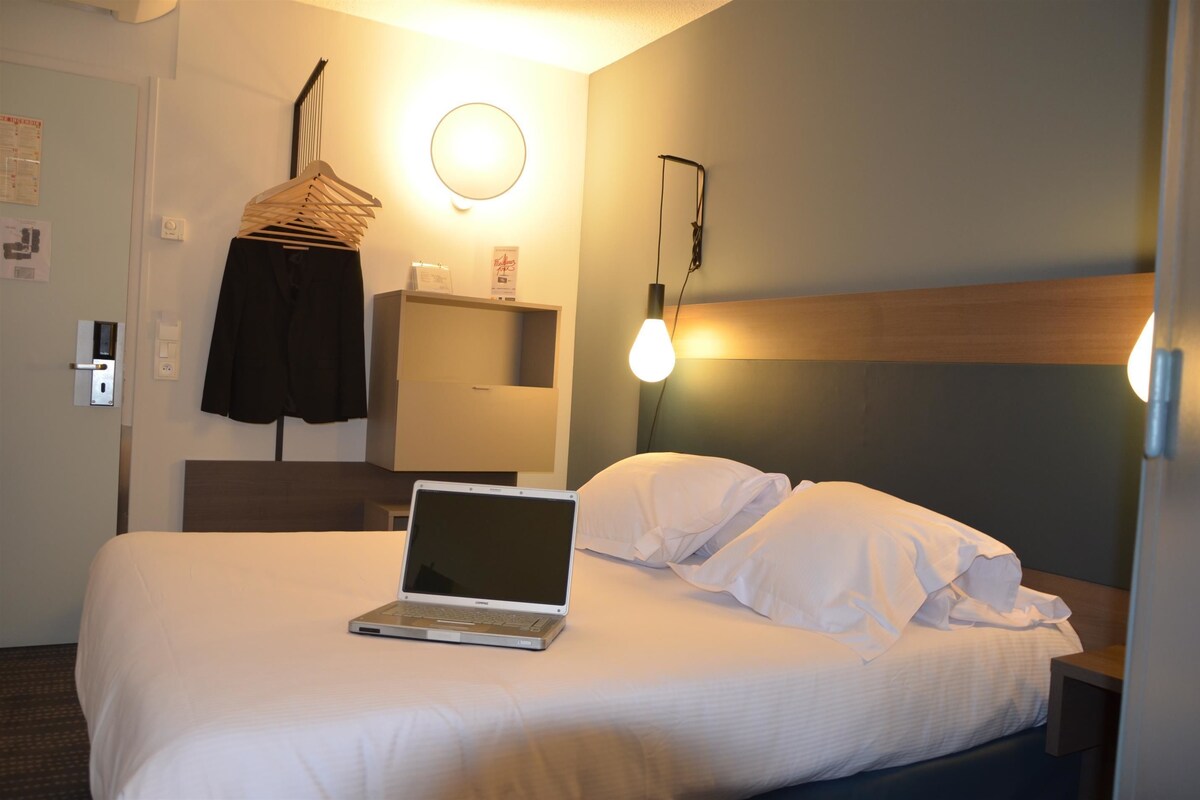 A hotel room with a bed, a laptop, a hanging rack, and a wall-mounted lamp.