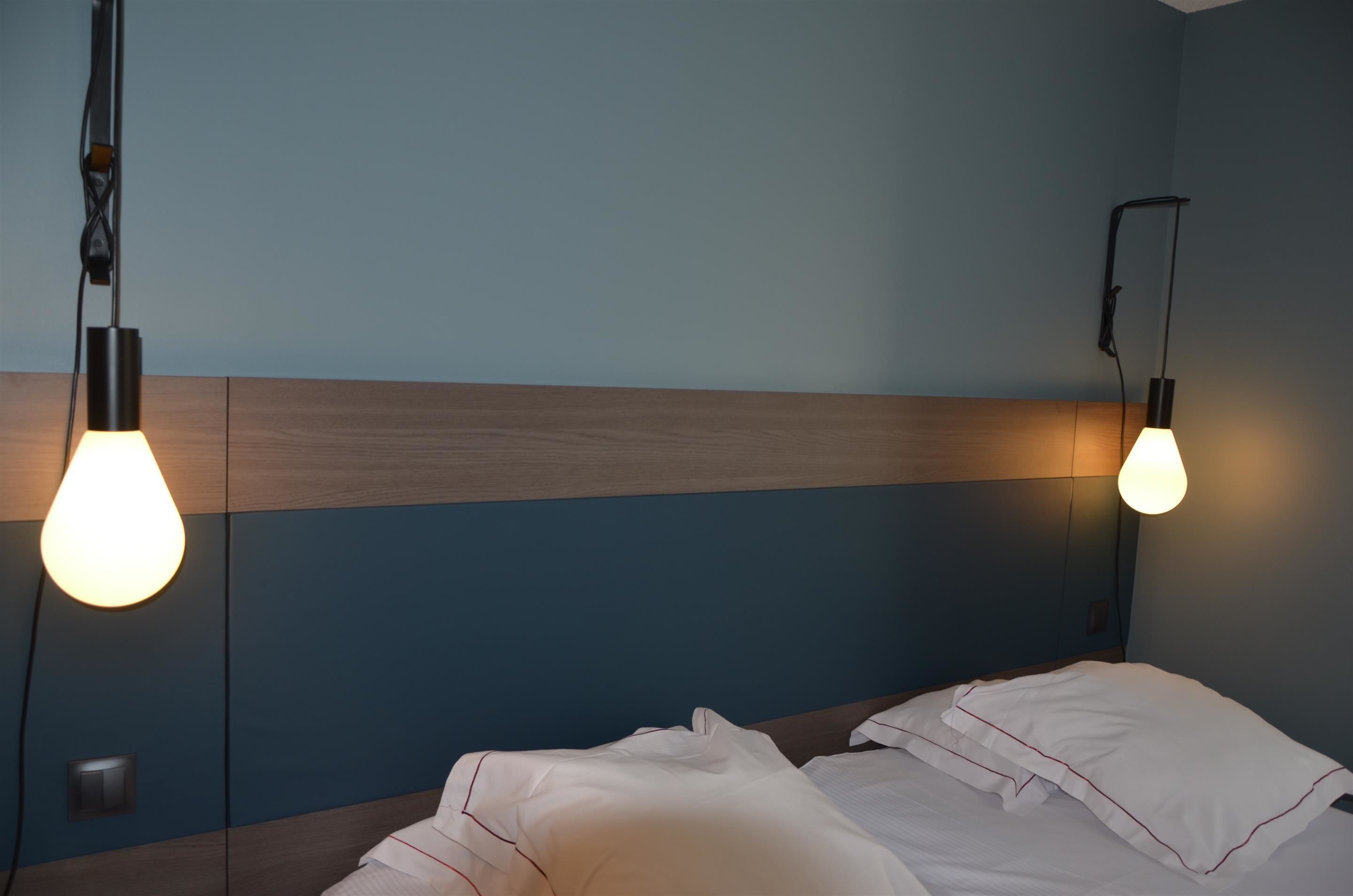 A hotel room with a bed, two wall-mounted light fixtures, and a wooden headboard.