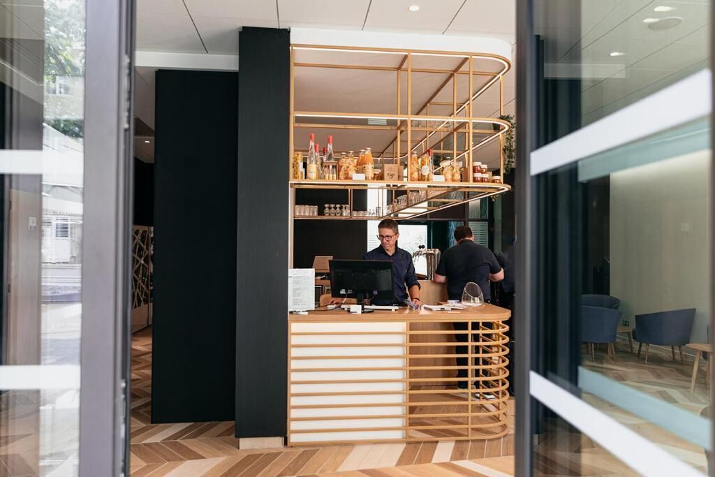A modern coffee shop interior with a wooden counter, shelves with various items, and a seating area visible through a glass door.