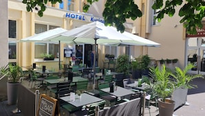 Een terras van een hotel met tafels en stoelen, waarvan een deel overdekt is met parasols, en omringd door potplanten.