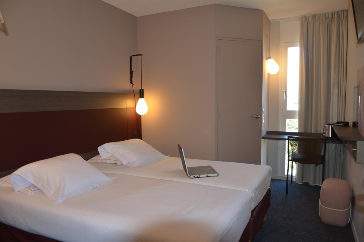 A hotel room with two beds, a desk, a laptop, a chair, and a window with curtains.