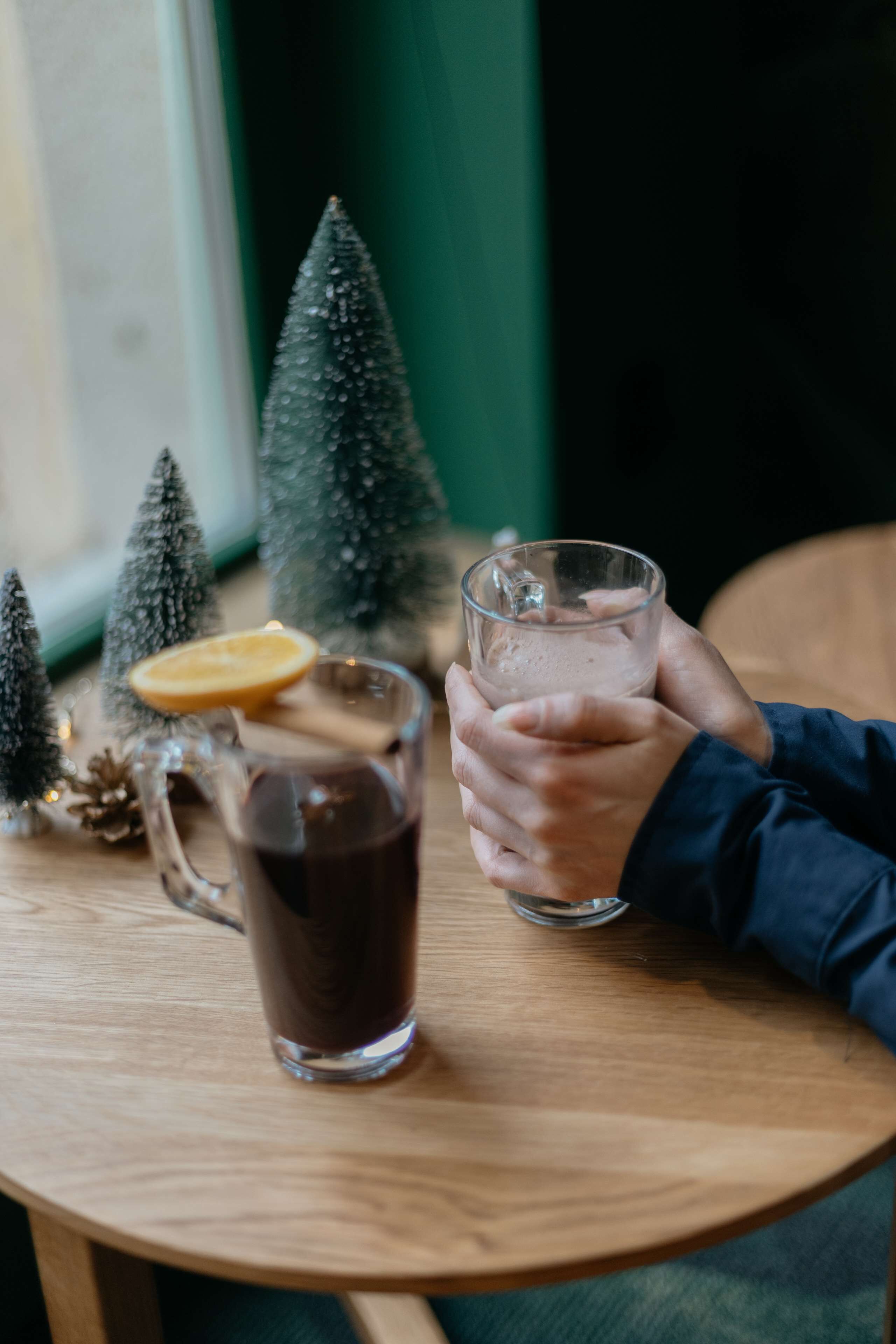 A wooden table with a glass mug and a glass of dark liquid, possibly tea or coffee, with a slice of orange and a cinnamon stick. There are small Christmas trees and decorations on the table.