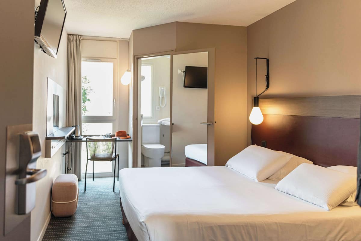 Habitación de hotel con cama, escritorio, silla, televisión y baño visible a través de una puerta abierta.