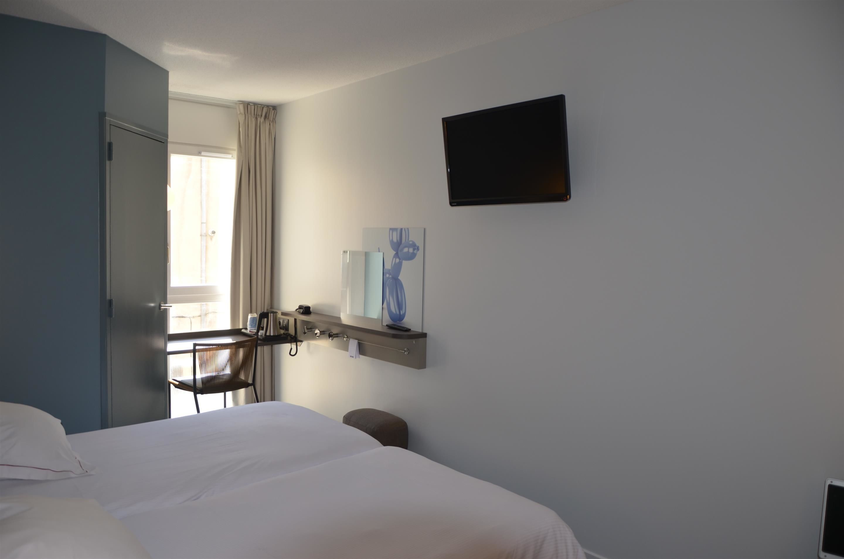Une chambre d’hôtel comprenant un lit, un bureau avec une chaise, un téléviseur fixé au mur et une fenêtre avec des rideaux.