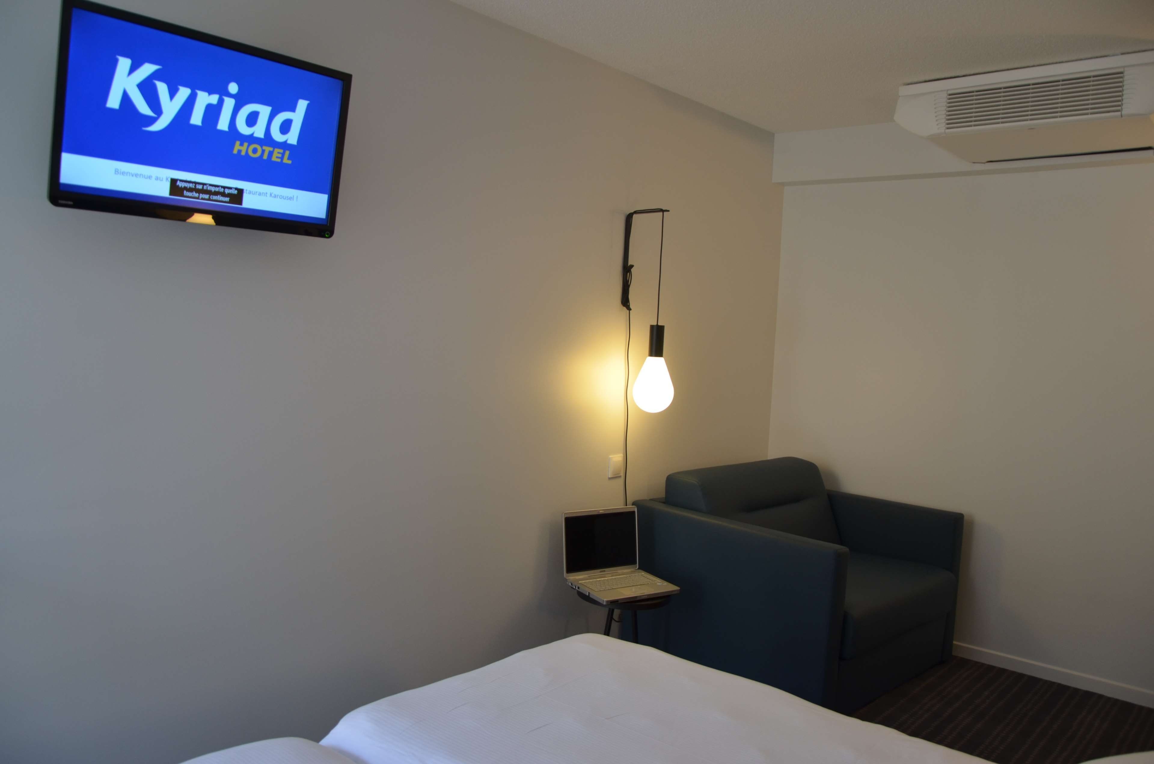 A hotel room with a bed, a desk with a laptop, a chair, a wall-mounted TV displaying 'Kyriad HOTEL', and an air conditioning unit.