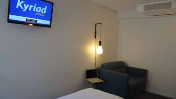 A hotel room with a bed, a desk with a laptop, a chair, a wall-mounted TV displaying 'Kyriad HOTEL', and an air conditioning unit.