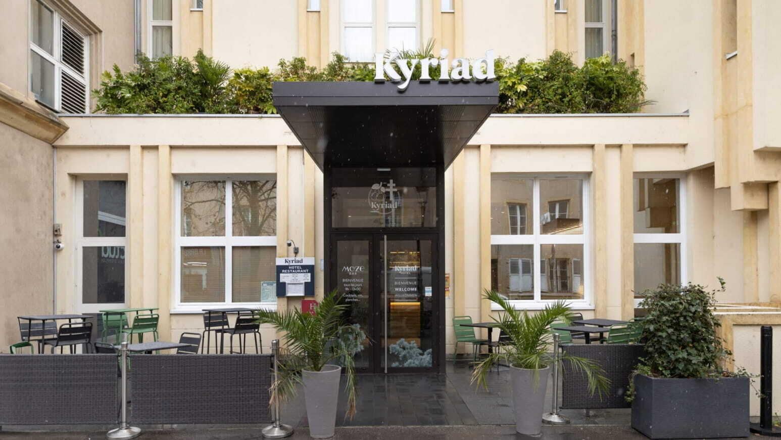 A hotel entrance with a sign reading 'Kyriad' and outdoor seating with tables and chairs.