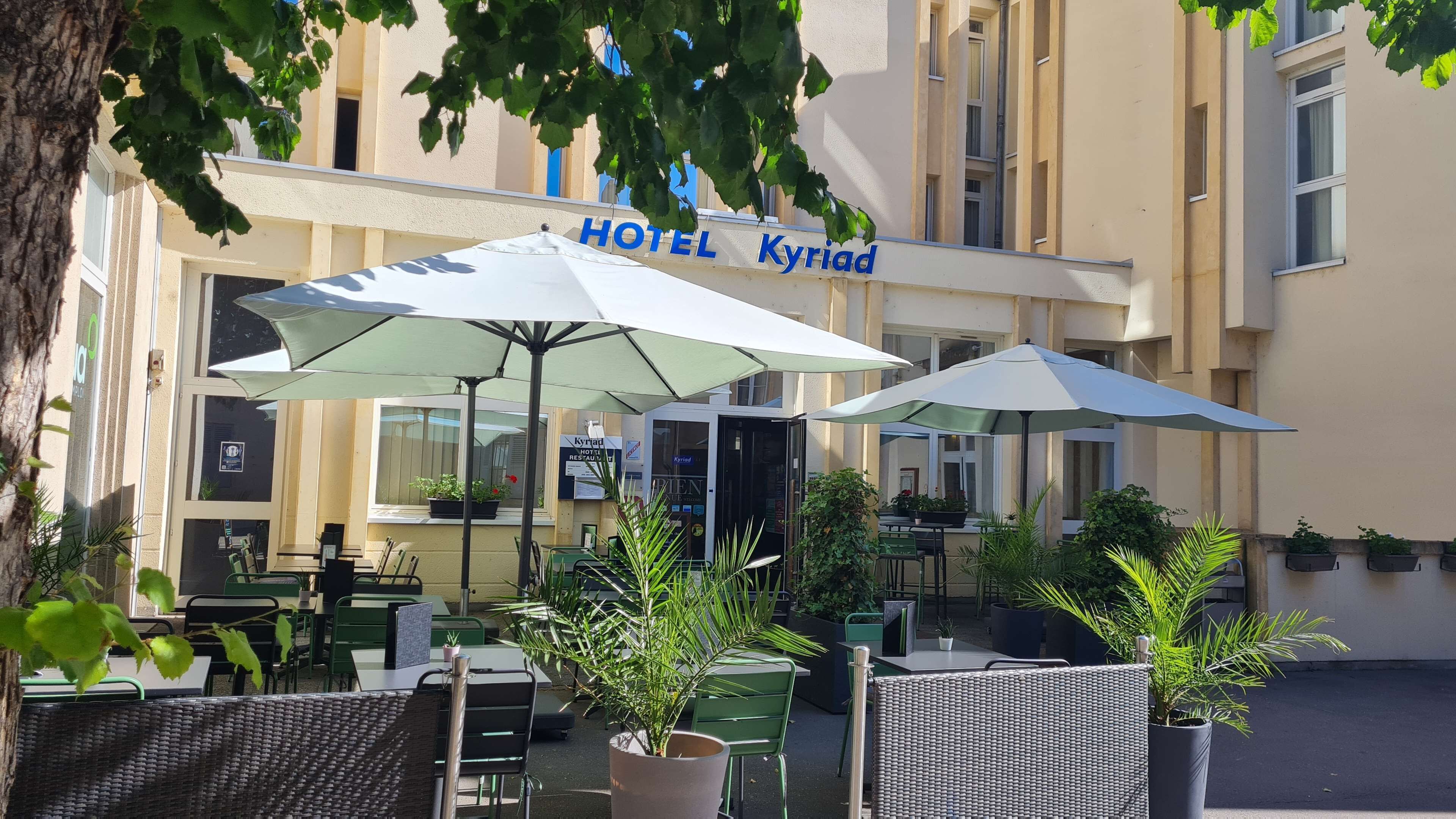 A hotel exterior with outdoor seating, umbrellas, and potted plants.