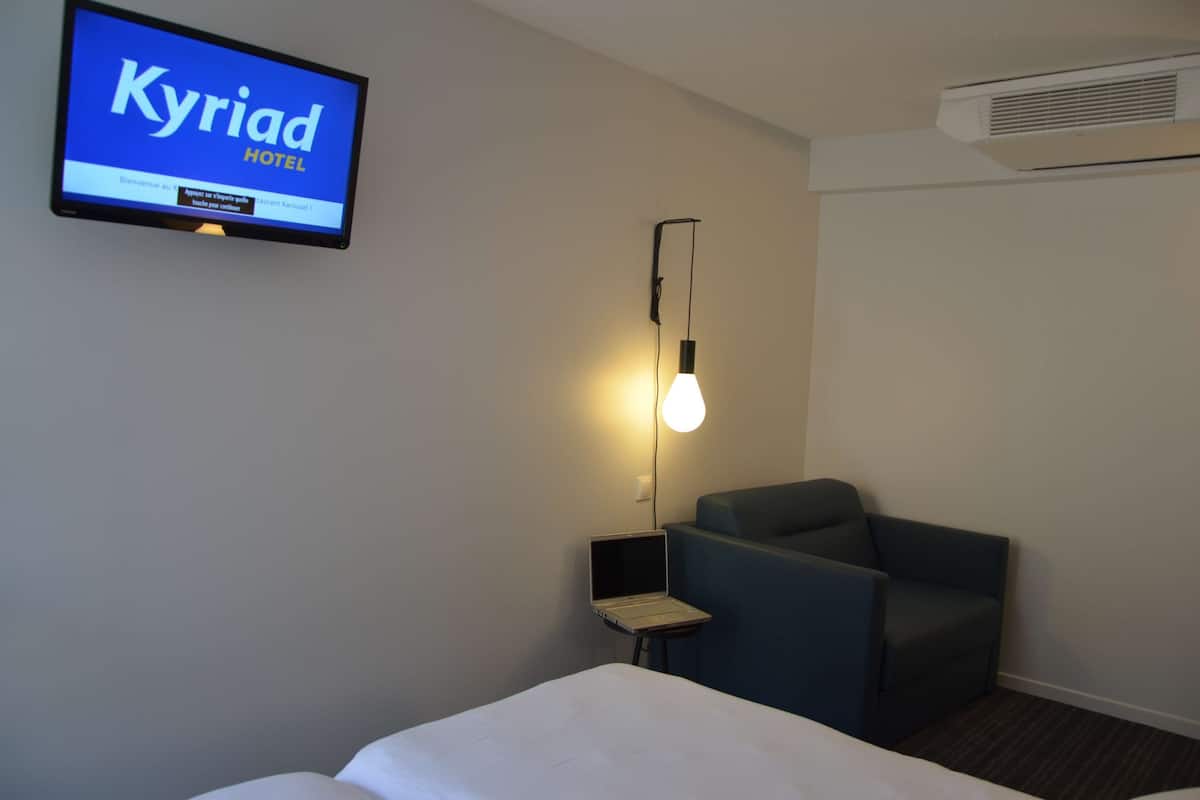Una habitación de hotel con una cama, un escritorio con una computadora portátil, una silla, un televisor empotrado que muestra 'Kyriad Hotel' y un aire acondicionado.