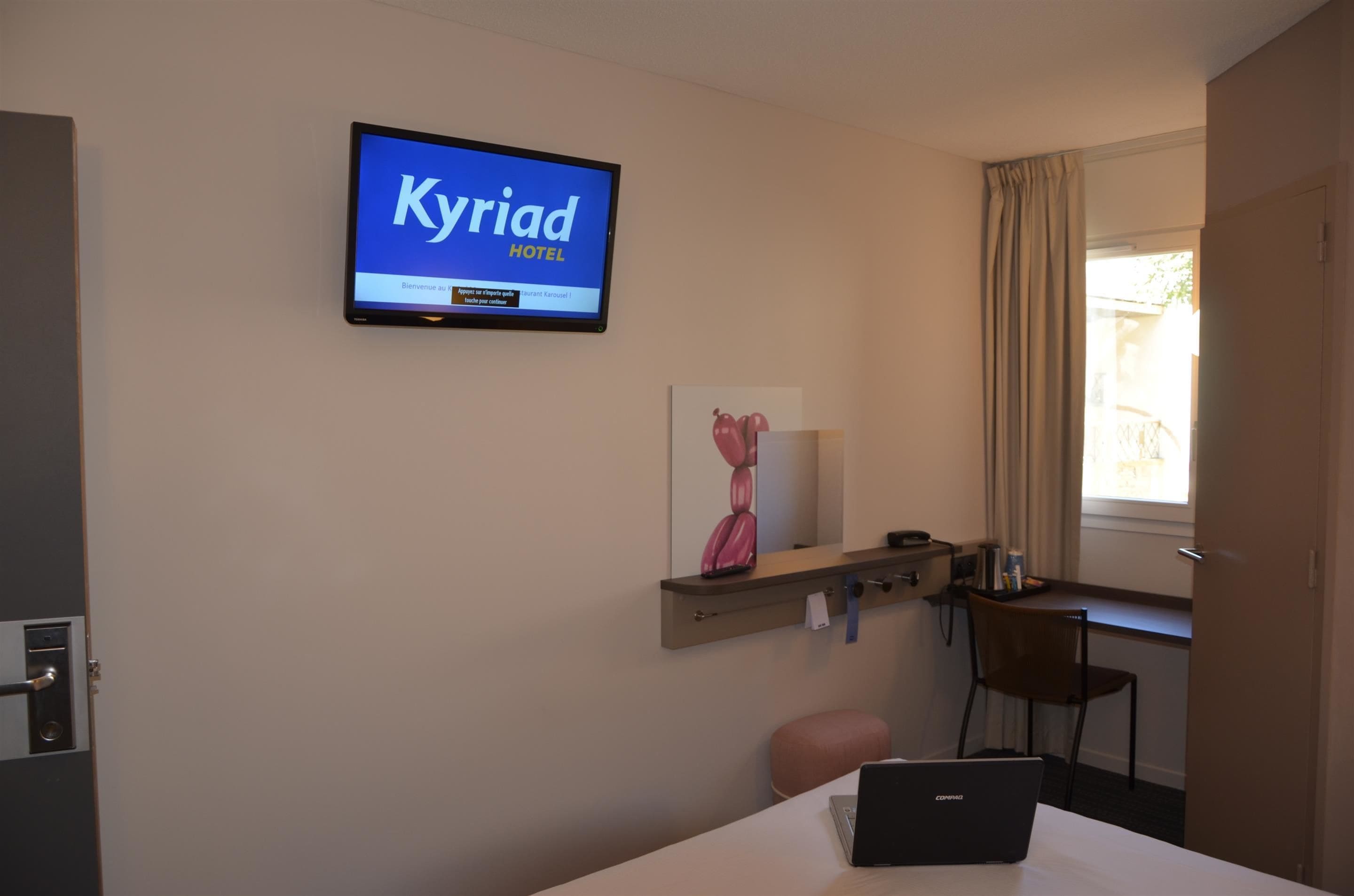 Una habitación de hotel con una televisión mostrando 'Kyriad Hotel' en la pared, un escritorio con una computadora portátil, una silla y una pequeña mesa con un florero.