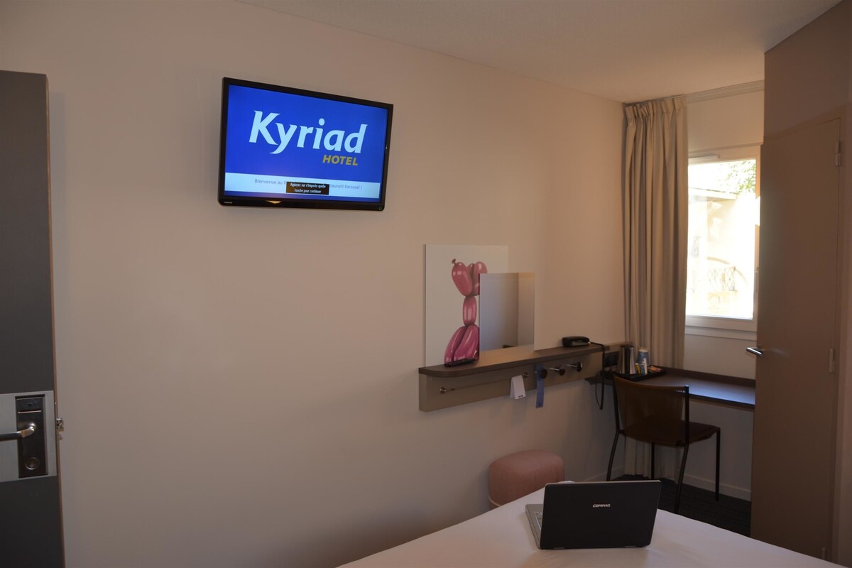 A hotel room with a television displaying 'Kyriad Hotel' on the wall, a desk with a laptop, a chair, and a small table with a vase.