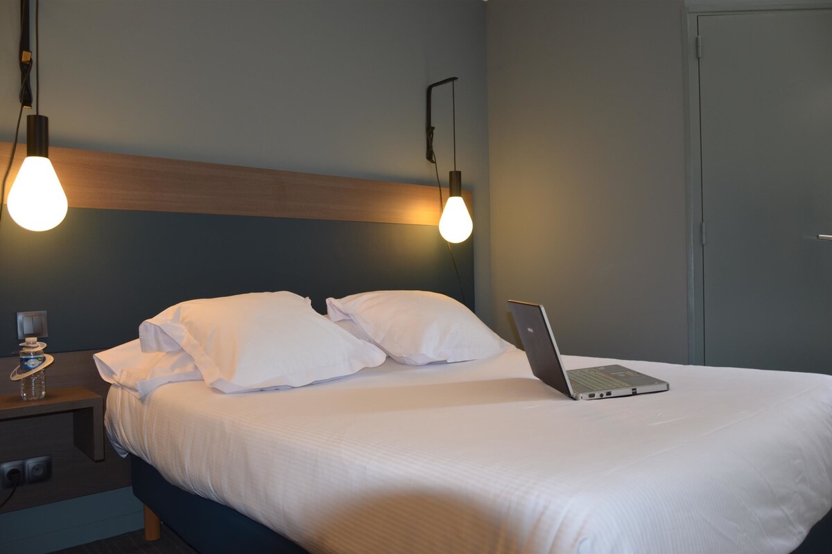 A neatly made bed with white linens and pillows, a laptop on the bed, and two pendant lights hanging above.