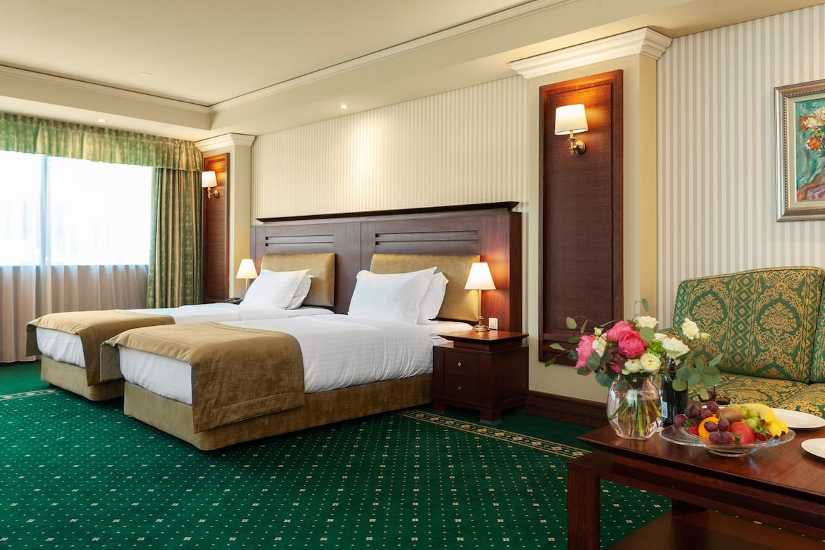 A hotel room with two beds, a sofa, a dining table with fruit and flowers, and a painting on the wall.
