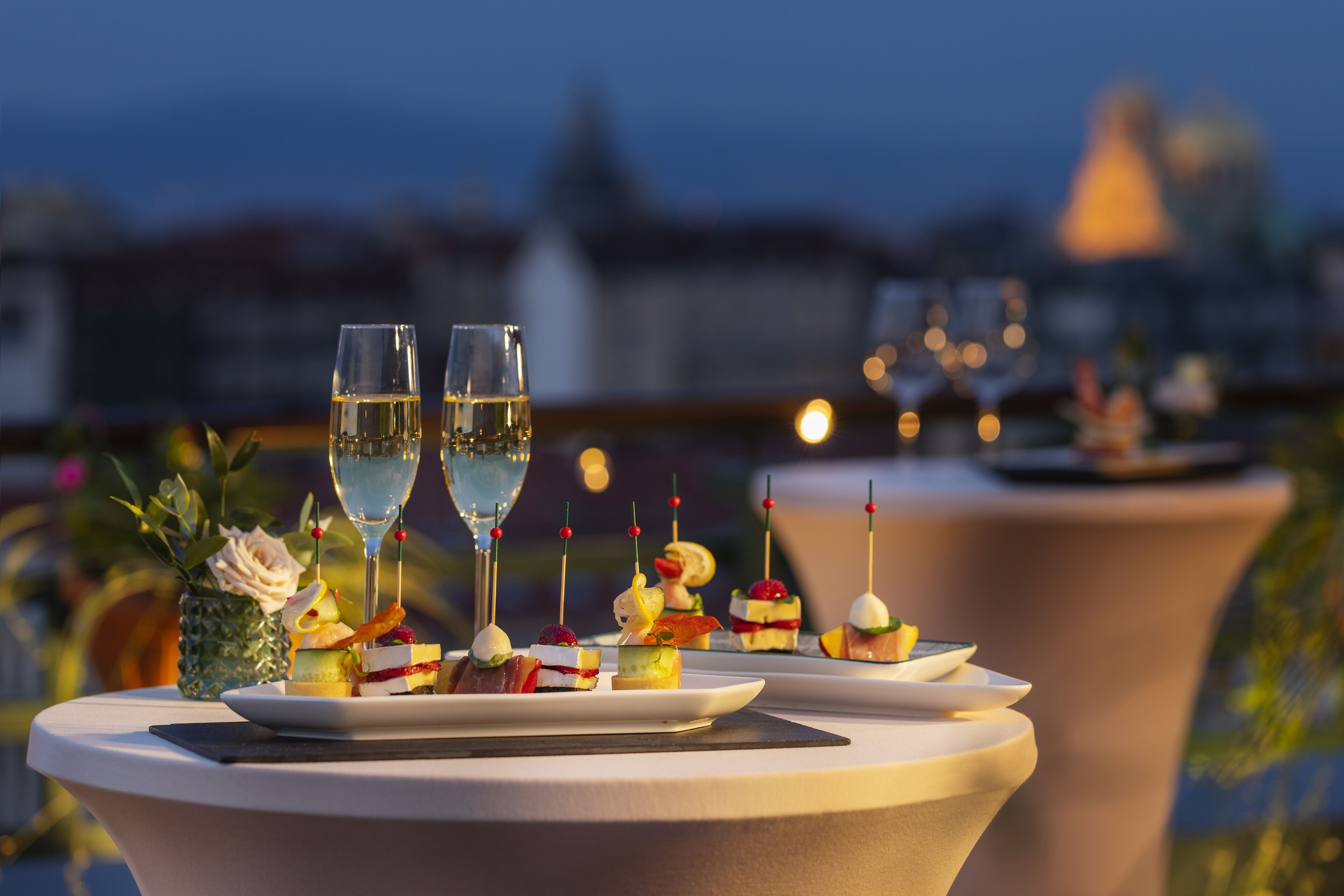 A table with a variety of appetizers and two glasses of champagne, set against a cityscape backdrop.