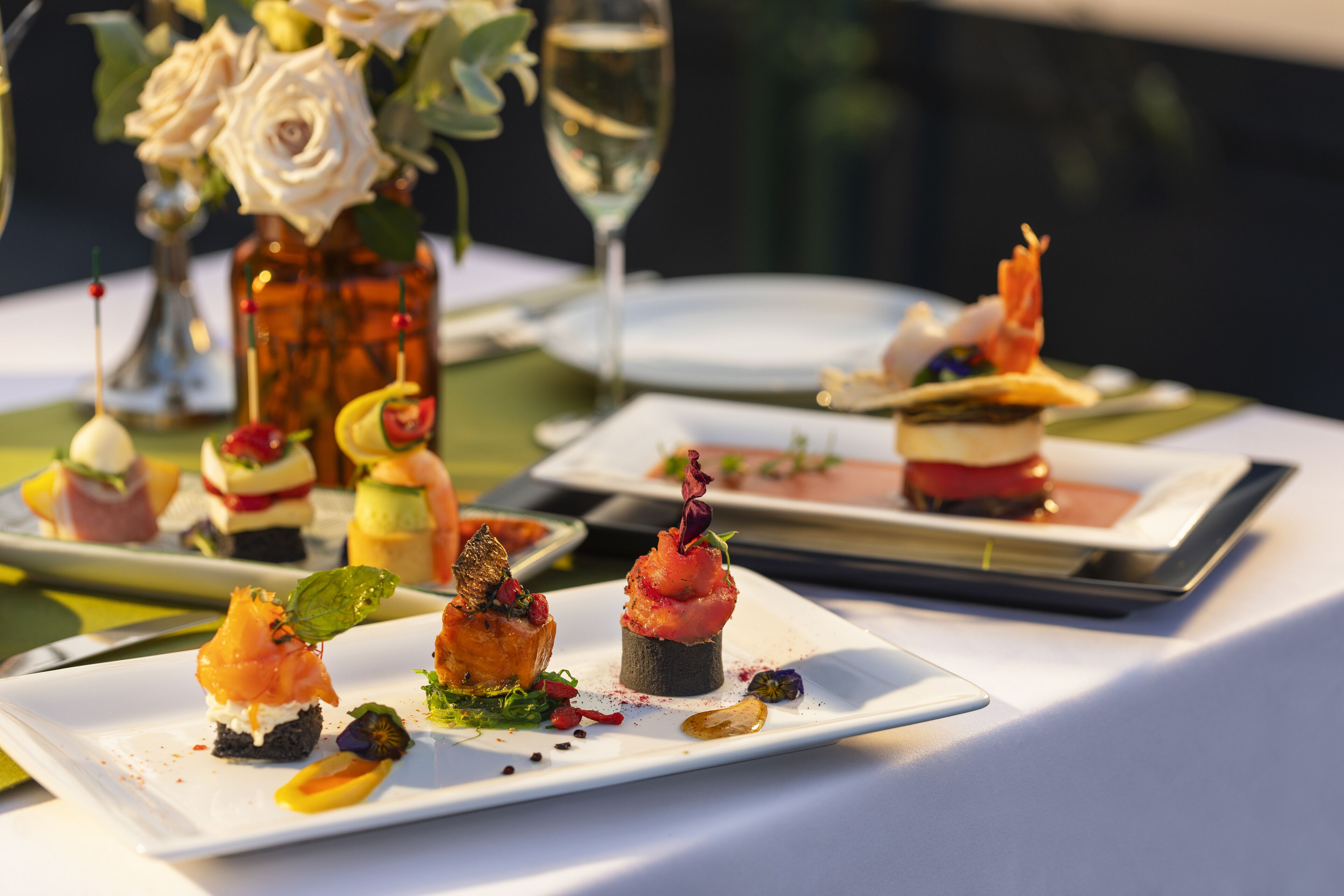 Ein Tisch ist reichhaltig mit Gourmet-Vorspeisen, einem Glas Weißwein und einer Vase mit weißen Blumen gedeckt.
