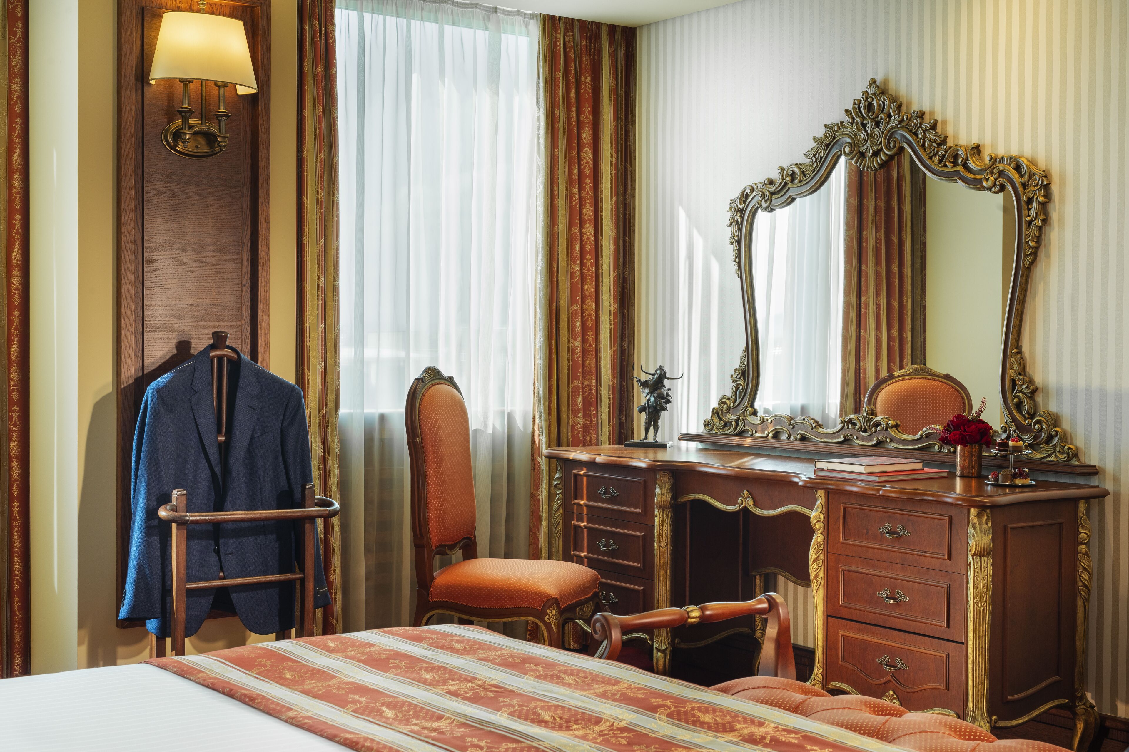 A hotel room with a bed, a wooden dresser with drawers, a mirror, a chair, and a coat rack with a jacket.