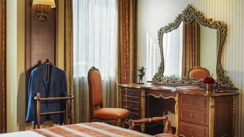 A hotel room with a bed, a wooden dresser with drawers, a mirror, a chair, and a coat rack with a jacket.