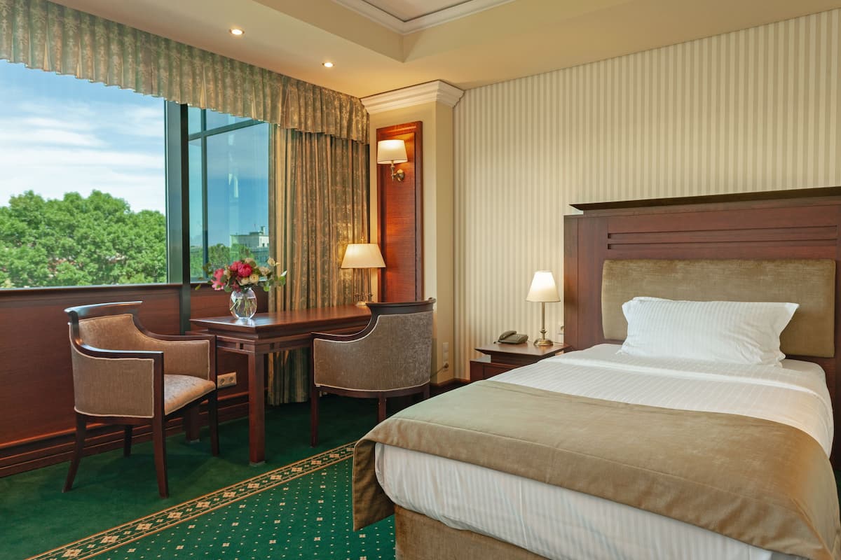A hotel room with a large bed, two chairs, a desk, and a view of greenery outside.