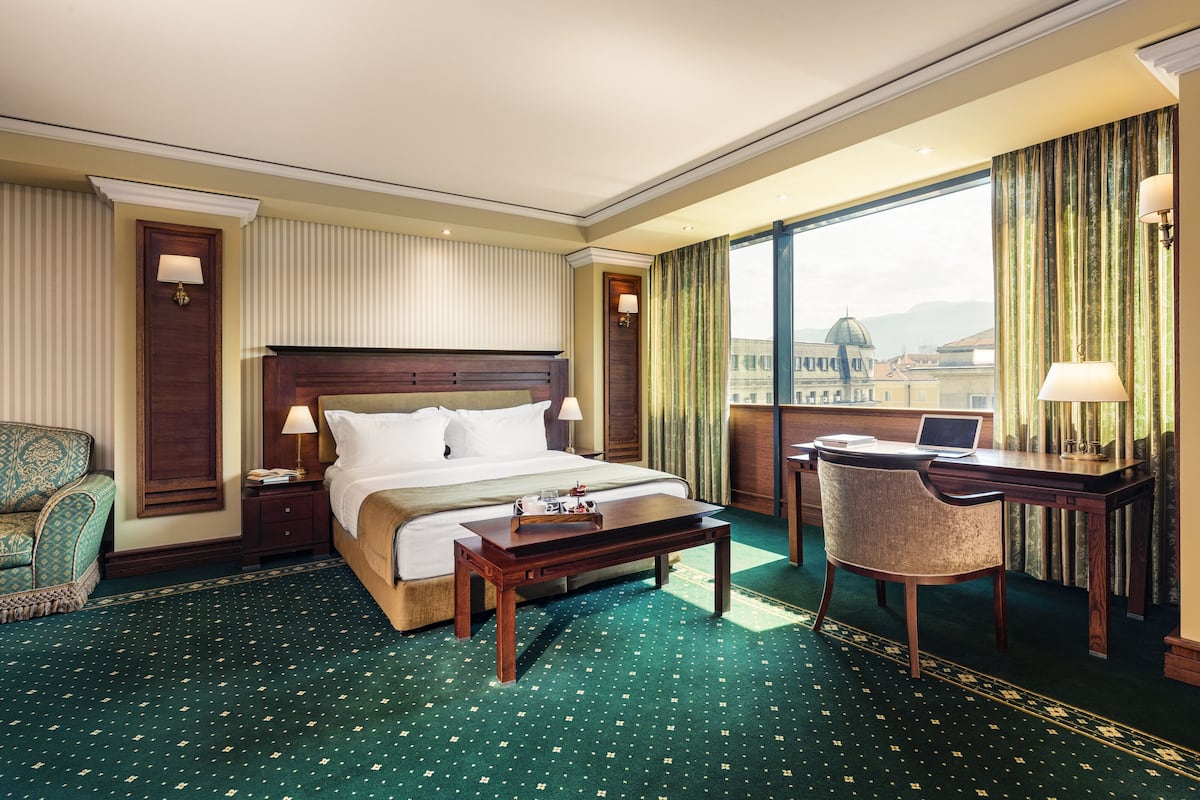 A hotel room with a large bed, a desk with a laptop, a chair, and a view of a cityscape through large windows.