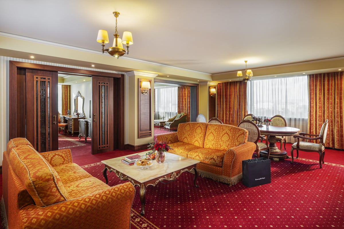 A spacious hotel room with a red carpet, a sofa, a coffee table with a vase of flowers, and a dining area with a table and chairs.