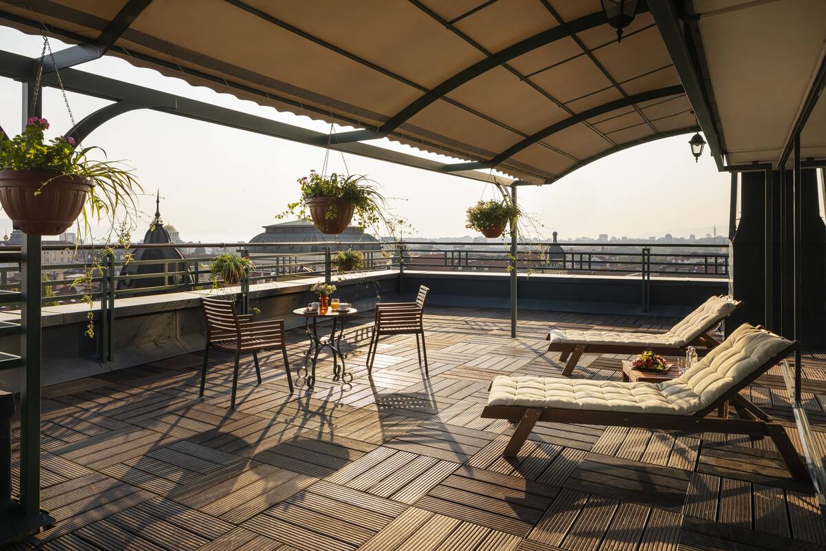A rooftop terrace with wooden flooring, two lounge chairs, a table with chairs, hanging planters, and a view of a cityscape.