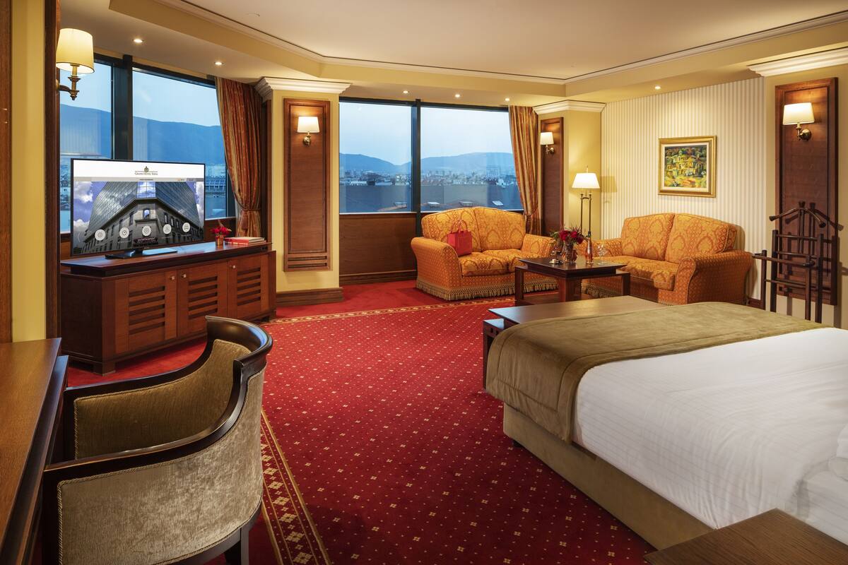 A hotel room with a large bed, a desk with a computer, a sofa, and a view of a cityscape through large windows.