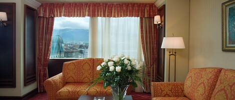 A hotel room with two patterned sofas, a coffee table with a vase of flowers, a lamp, and a view of a cityscape through large windows.