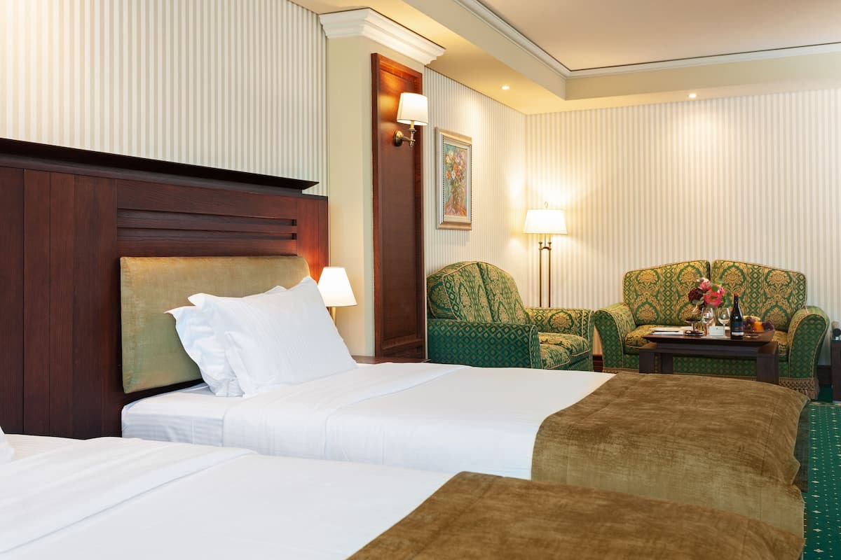 A hotel room with two beds, a green patterned armchair, a wooden headboard, and a small table with a vase and bottle.