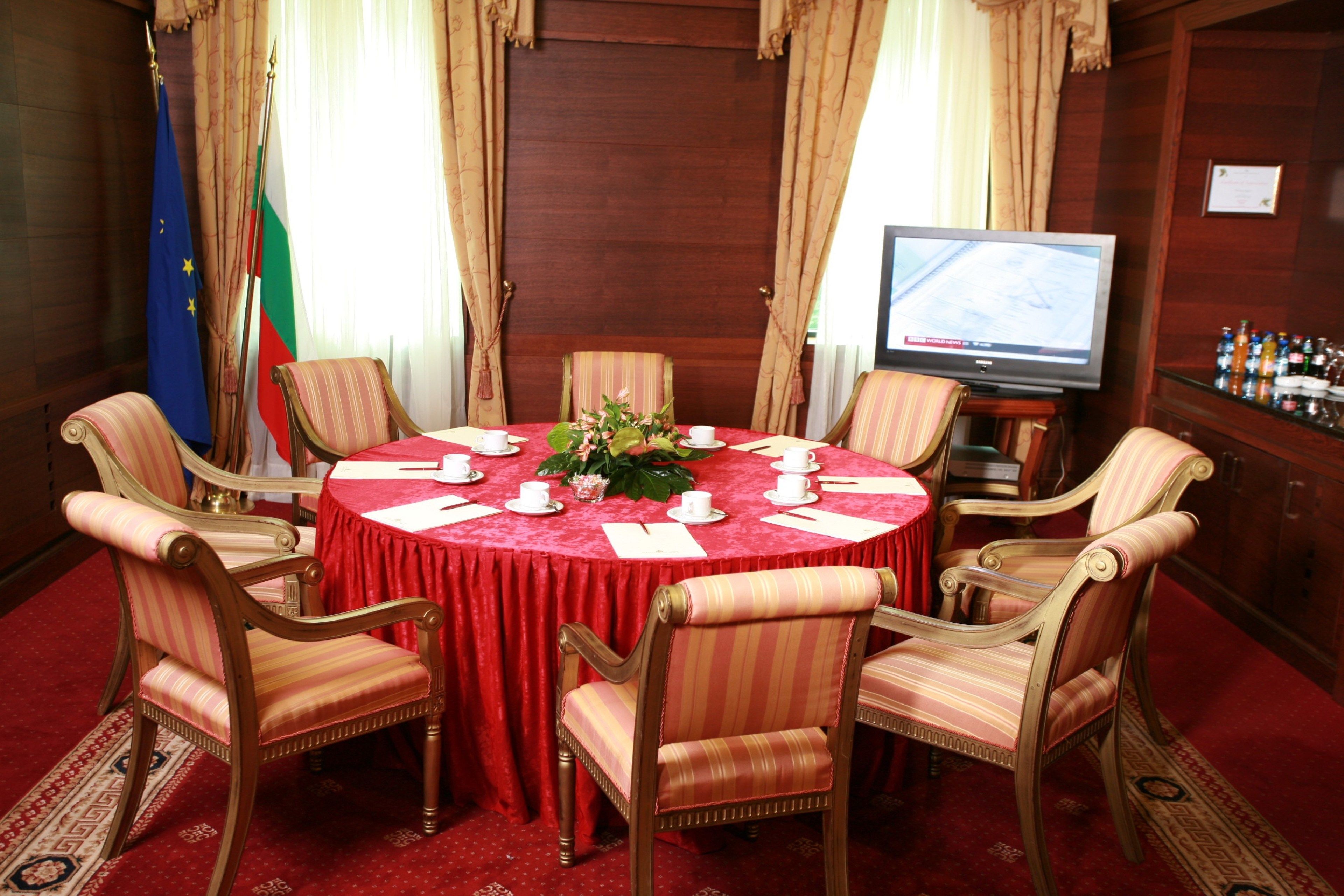 Un salón de conferencias con una mesa redonda, sillas, banderas, un televisor y una zona de bar.