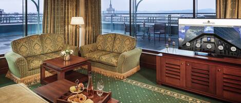 Habitación de hotel con sofá de color verde estampado, mesa de centro de madera con bandeja de aperitivos y bebidas, lámpara y un ventanal que ofrece vistas a un paisaje urbano y un puente.