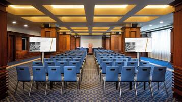 A conference room with rows of blue chairs, a patterned carpet, wooden paneling, and two screens displaying conference room information.