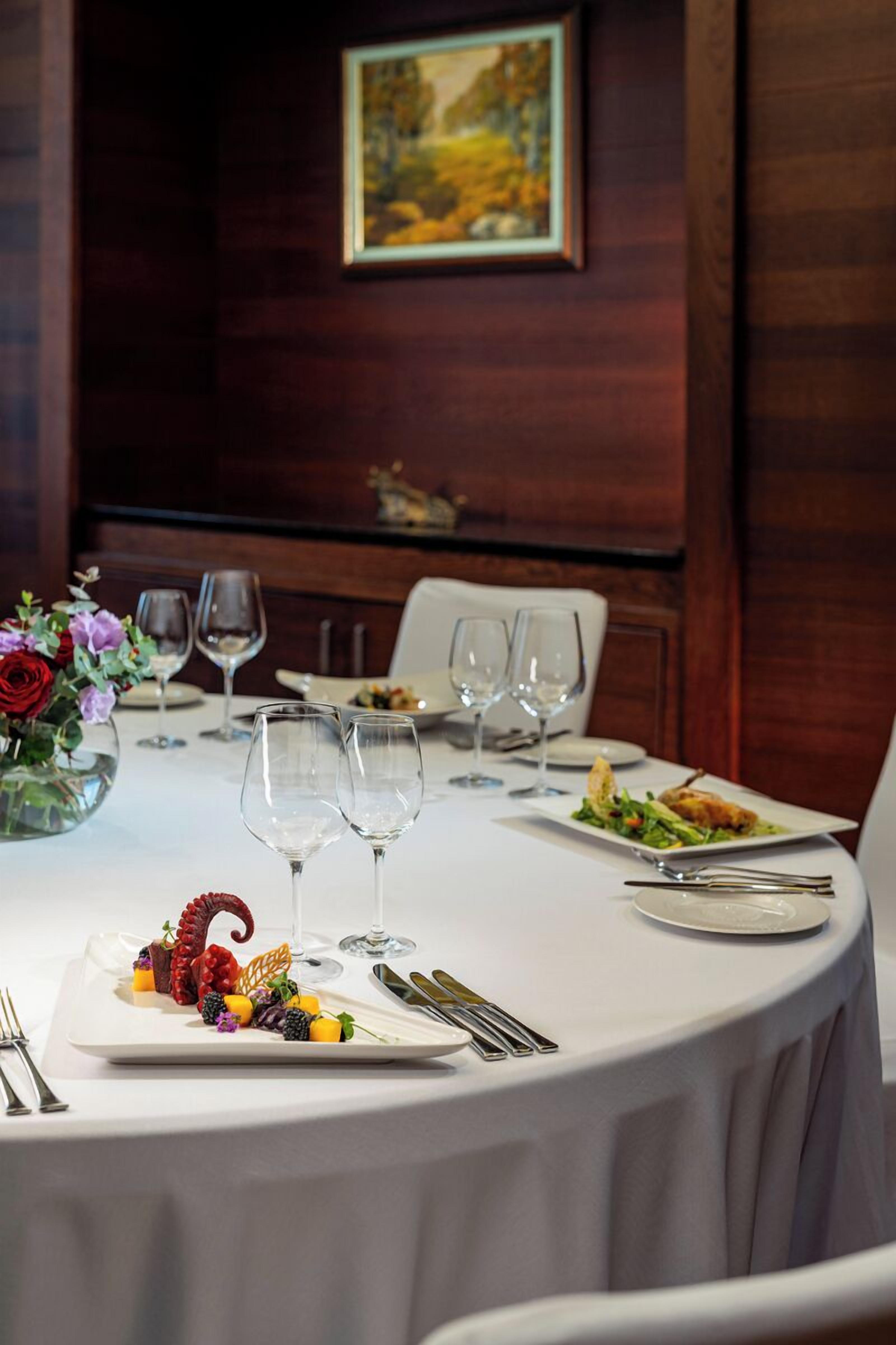 Una mesa redonda para comer, con mantel blanco, platos, cubiertos y copas de vino. Un jarrón con flores está sobre la mesa.