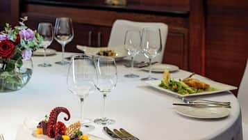 Una mesa redonda para comer, con mantel blanco, platos, cubiertos y copas de vino. Un jarrón con flores está sobre la mesa.