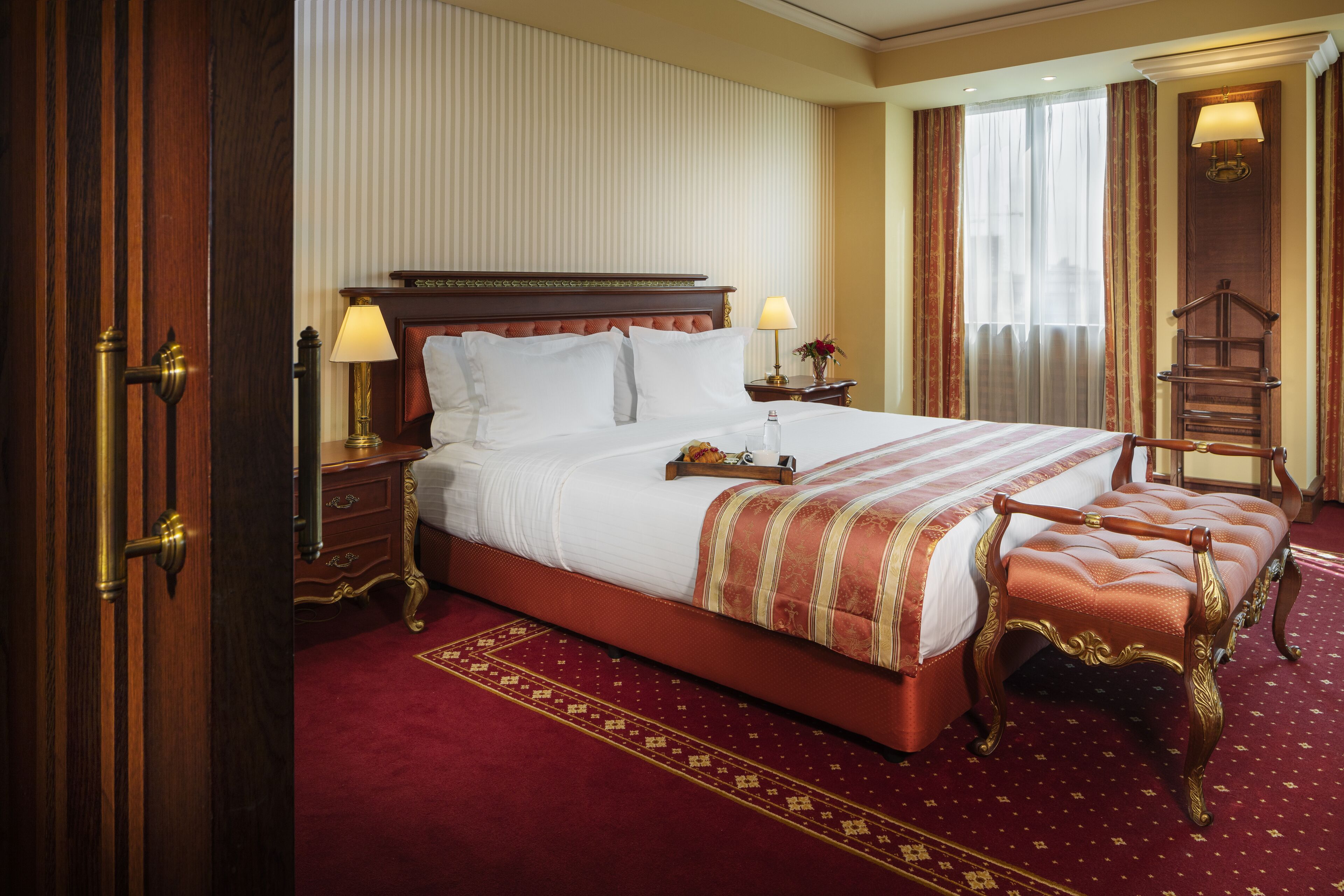 A large bed with a wooden headboard, two bedside tables with lamps, a bench, and a red carpeted floor.