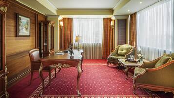 Habitación de hotel con pared de paneles de madera, alfombra roja, mesa de comedor, sofá y un gran ventanal con cortinas.
