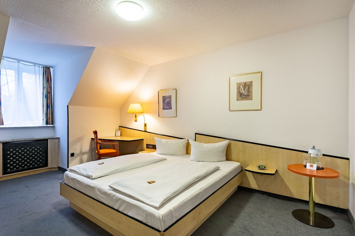 Una camera d'albergo con un letto grande, un comodino, una sedia, una lampada e un tavolino con una bottiglia.
